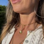 Layered gold charm necklaces on a woman in sunlight, styled with a white crochet blouse.