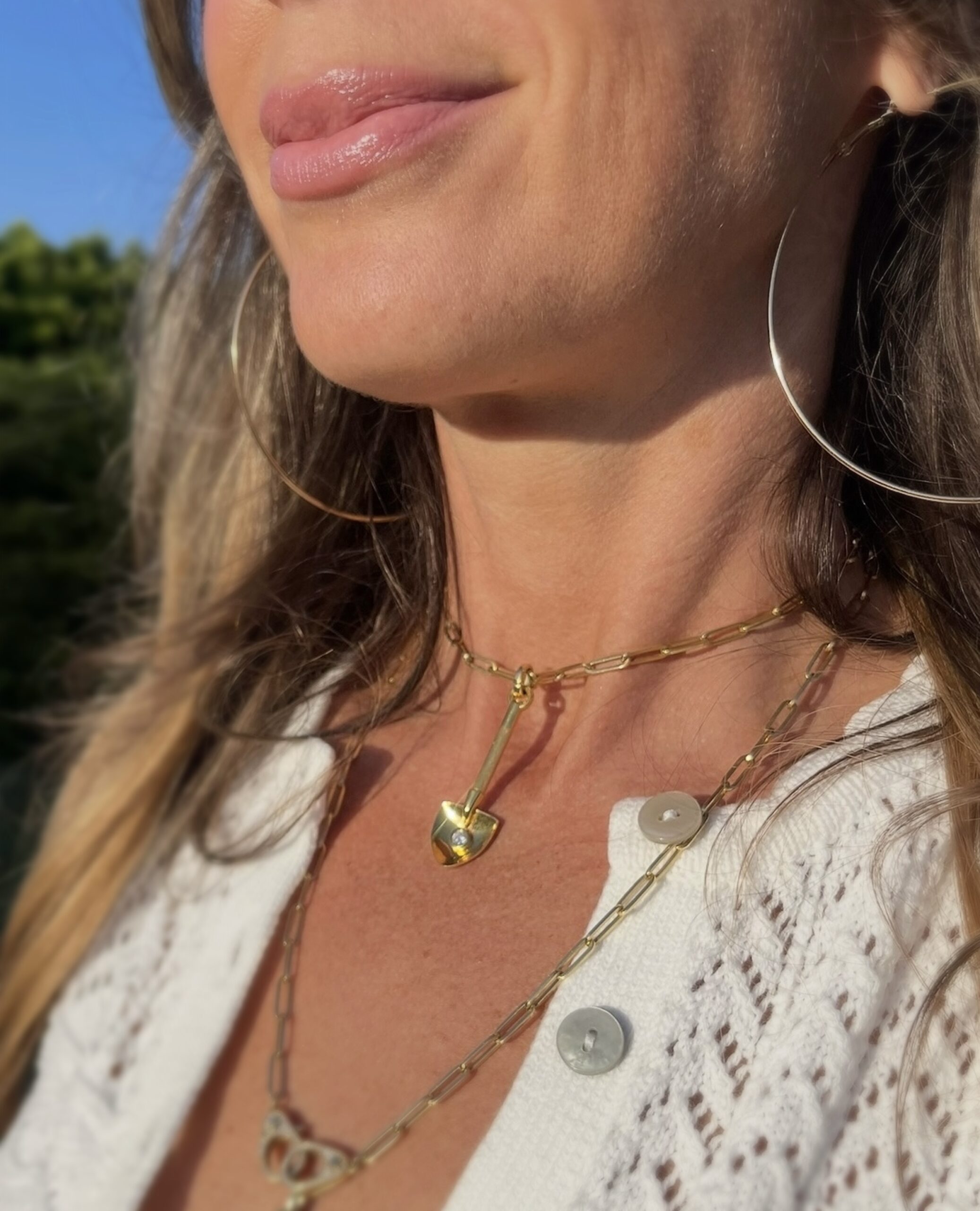 Layered good shovel diamond charm in summer light Layered gold charm necklaces on a woman in sunlight, styled with a white crochet blouse.